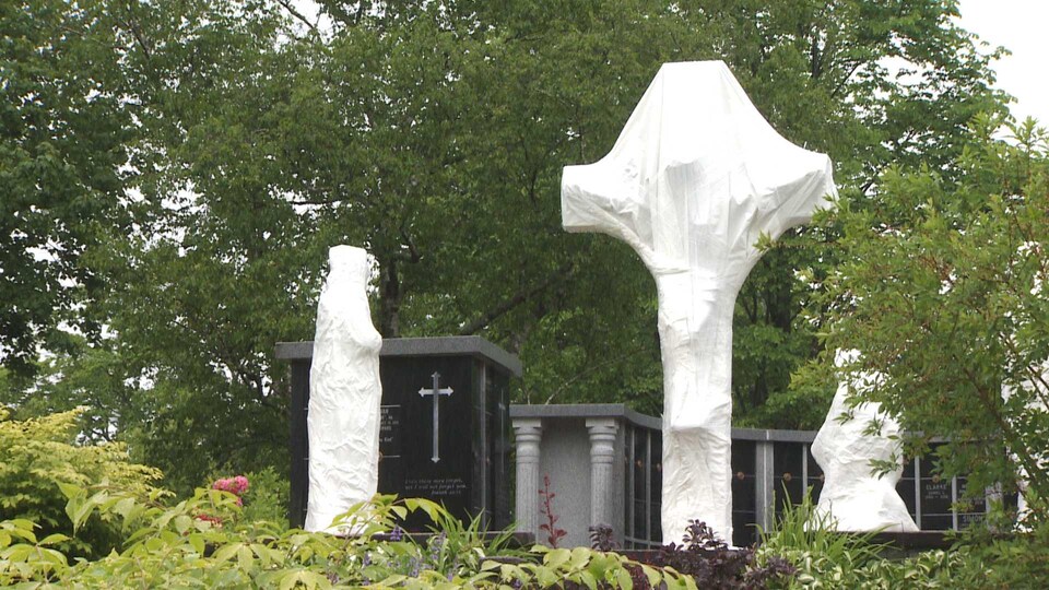 Une croix et des statues dans un cimetière sont recouverts de bâches blanches.