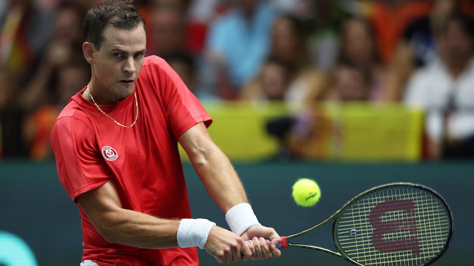 A tennis player, dressed in red and white, hits a backhand ball with both hands during a Davis Cup match. 