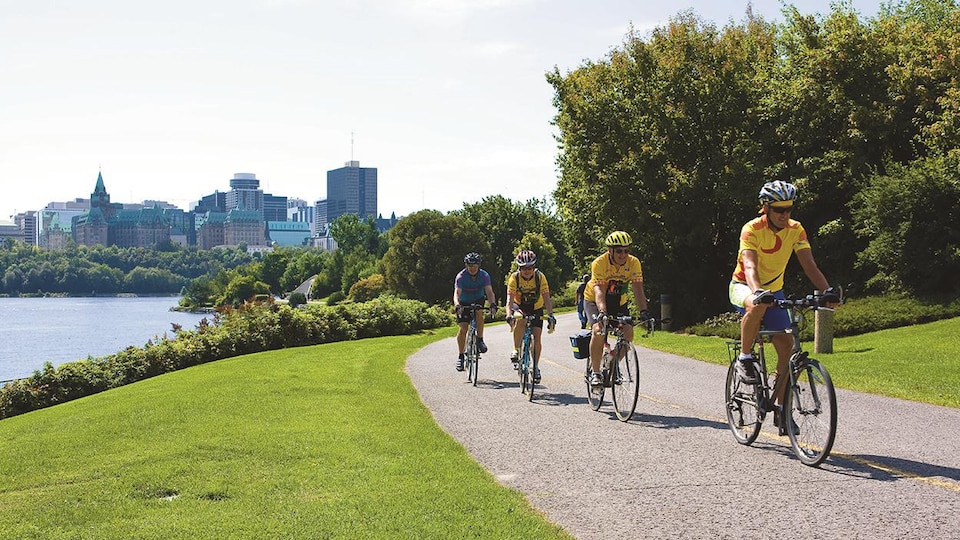 Des cycliste sur une piste cyclable en bordure de la rivière des Outaouais. Ottawa en arrière-plan.