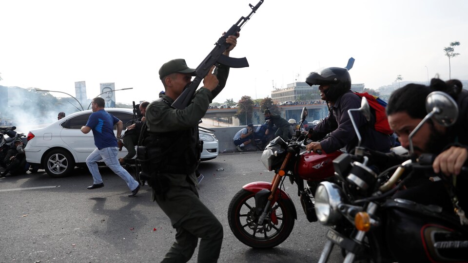 Un soldat pointe son arme dans les airs dans une rue où des civils courent et des hommes se déplacent à moto.