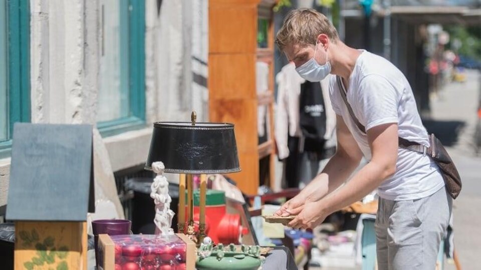 Un homme masqué se penche sur une table installée à l'extrieur et sur laqelle sont disposés beaucoup d'objets de décoration et anciens. Il tient une assiette dans ses mains et la regarde.