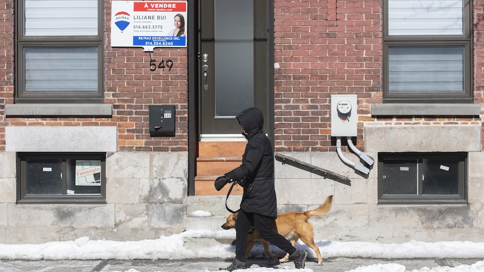 Une passante et son chien devant une maison à vendre, l'hiver.