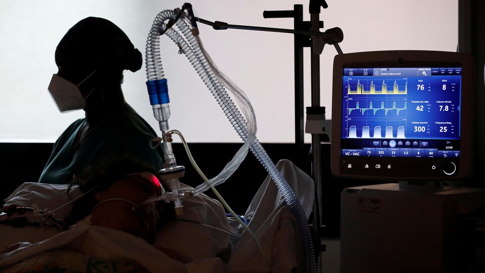 La silhouette d'une infirmière de profil se dessine sur un mur blanc. Elle est assise auprès d'une personne qu'on ne peut pas distinguer, mais qui est couchée sur un lit branchée à un respirateur artificiel et à des machines.