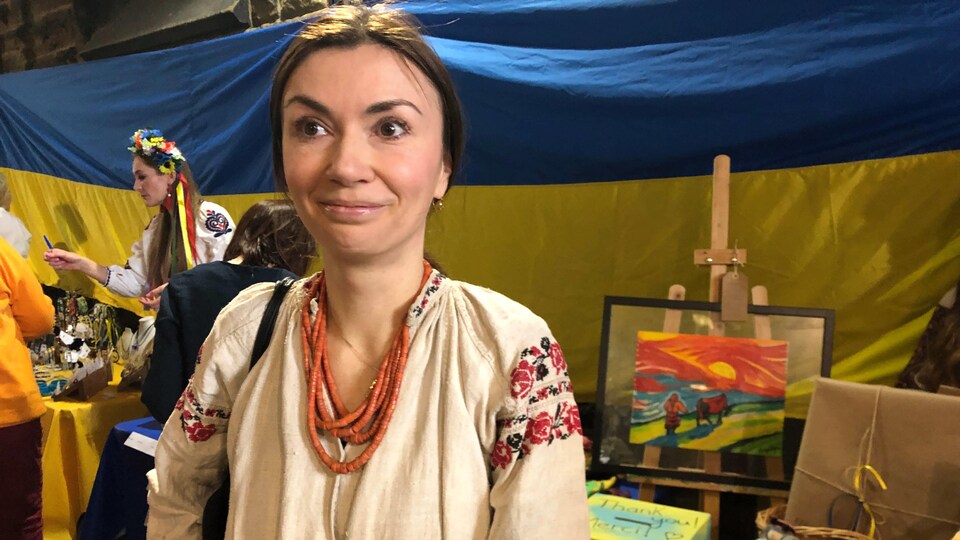 Vera Kostenko sourit pour une photo. Elle porte un habit traditionnel, est debout devant des tables remplies d'objet à vendre et de grands drapeaux ukrainiens bleus et jaunes accrochés au mur.