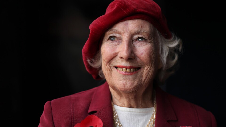 Portant un béret rouge et un veston rouge sur lequel sont attachés un coquelicot et ce qui ressemble à une insigne militaire, Dame Vera Lynn prend la pose pour la caméra.