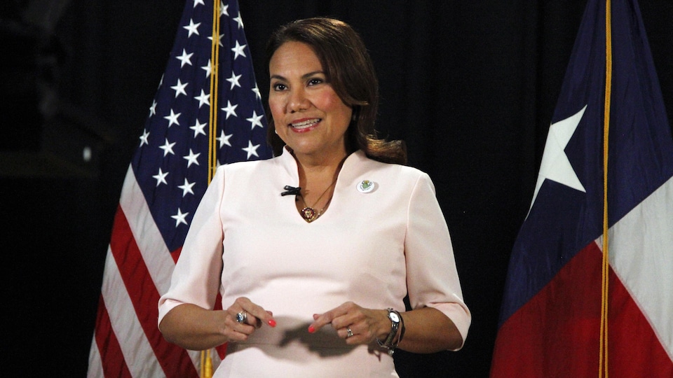 Une femme livre un discours. En arrière-plan, on retrouve un drapeau des États-Unis et un du Texas.