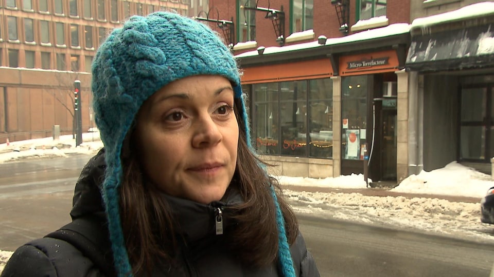 Véronique Lalande accorde une entrevue sur le trottoir situé en face de la station de Radio-Canada sur la rue Saint-Jean, à Québec. Elle porte une tuque de laine bleue et un manteau noir. La photo a été prise en hiver.