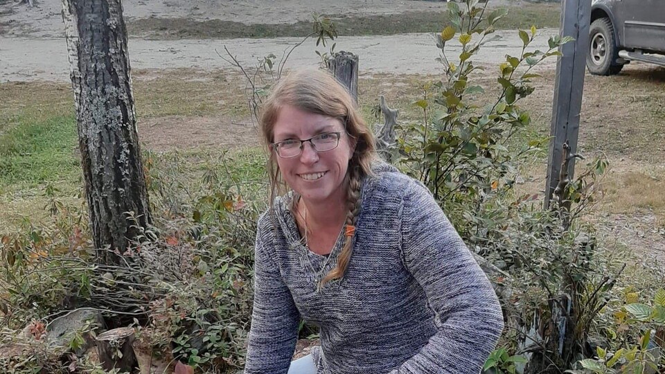 Le femme pose près d'une petite forêt. Elle sourit et porte des lunettes.