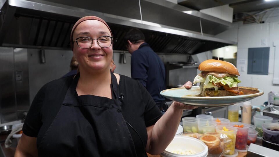Une cheffe dans sa cuisine tient dans sa main un burger de bœuf Wagyu avec de la salade, du. bacon et du fromage.