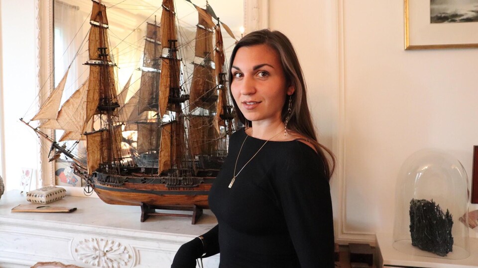 Joëlle Preston pose dans un espace intérieur, devant un foyer sur lequel trône une réplique de voilier.