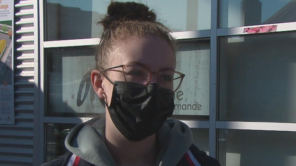 Une jeune femme portant des lunettes et un masque au visage, à l'extérieur d'un bâtiment public.