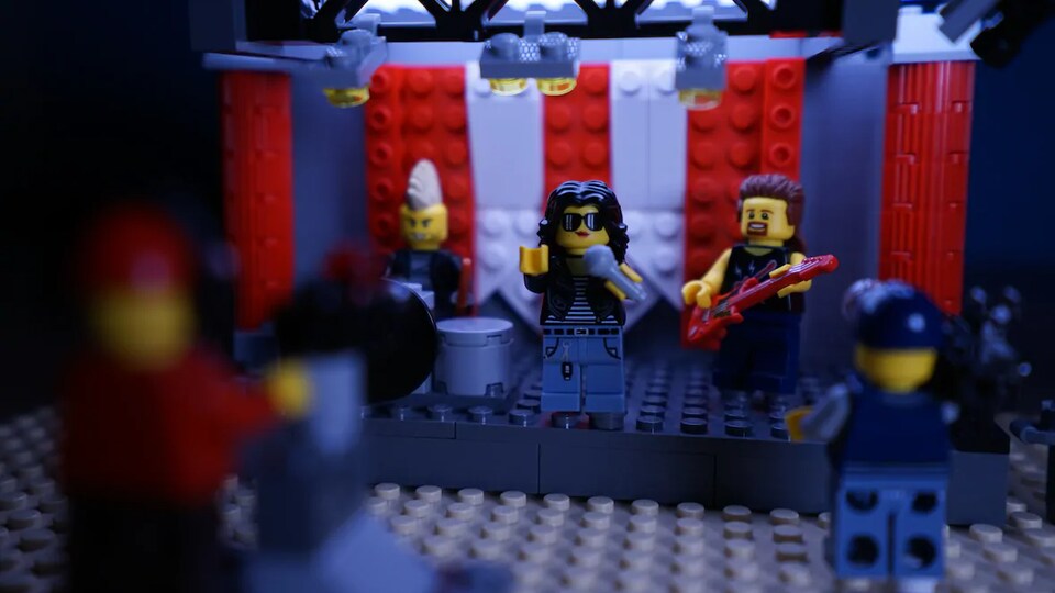 Des personnages Lego. Trois sont sur une scène : un qui joue de la guitare, un qui chante et un autre qui joue de la batterie. Devant eux deux autres personnages Lego filment.
