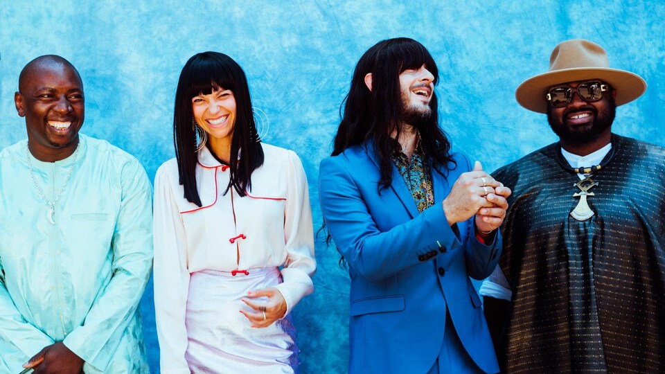 Les quatre musiciens posent tout sourire devant un mur bleu pâle. 