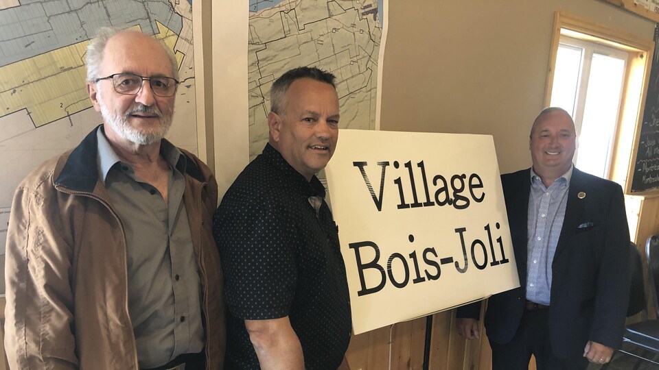 Trois hommes devant une affiche indiquant le nouveau nom: Village Bois-Joli
