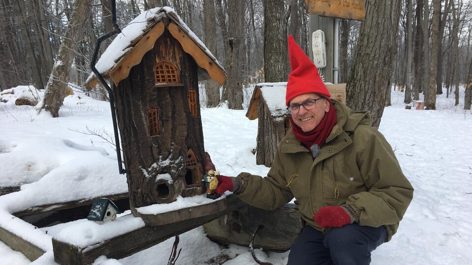 Un homme souriant s'agenouille pour poser avec un petit gnome et une bûche modifiée en petite maison.
