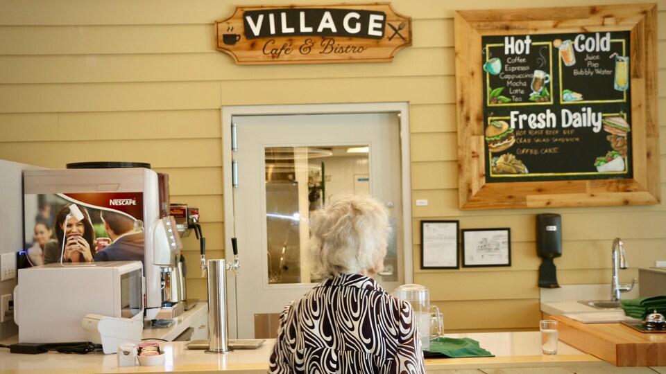 Une dame âgée, de dos, au comptoir d'un café tout en bois. 