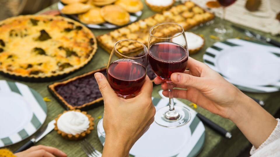 Deux personnes, dont on ne voit que les mains, trinquent avec leur verre de vin rouge. À l'arrière-plan, une table est remplie de nourriture. 