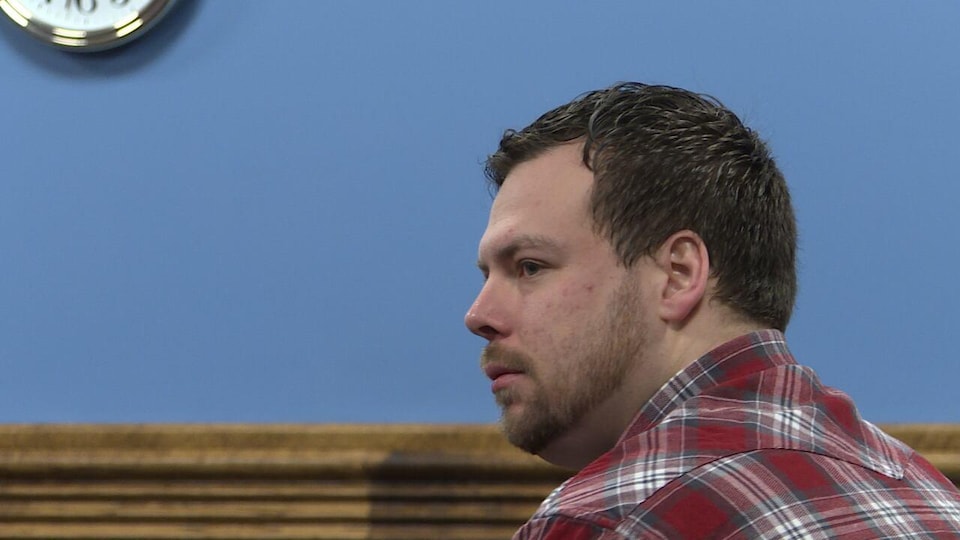 Un homme de profil assis dans une salle d'audience. Il porte une chemise à carreaux rouge.