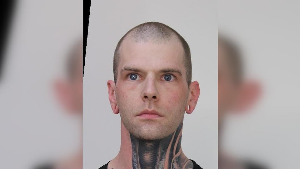 Un homme a la tête rasée et des tatouages sur le cou. Il a aussi les yeux bleus.