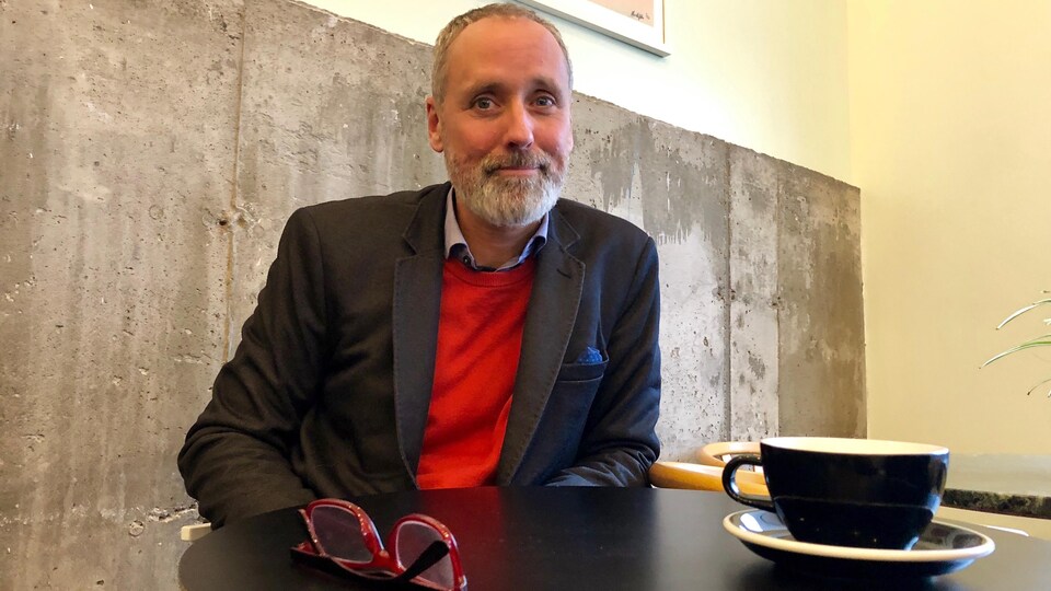Vincent Marissal assis à une table, où sont déposées des lunettes et un bol de café.