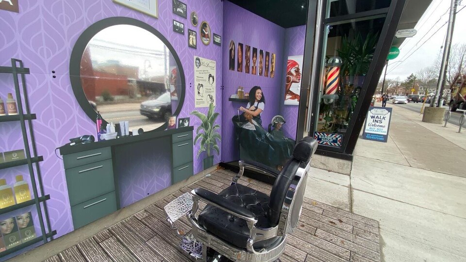 Une installation artistique extérieure. Une chaise de salon de coiffure est accessible aux passants de la rue. Sur les murs qui entourent la chaise, des murales autocollantes recréent le salon de coiffure de Viola Desmond. Par exemple, un autocollant d’une coiffeuse en train de laver les cheveux à une cliente se trouve sur le mur de droite. Il y a aussi, entre autres, un miroir, des produits de beauté sur une étagère, une plante tropicale.