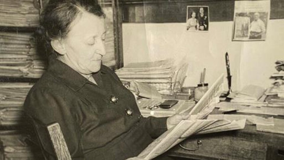 Assise sur une chaise, Violet McNaughton lit un journal.