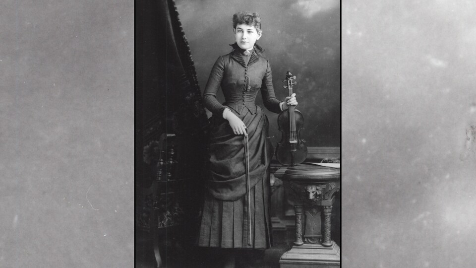 Elsie Reford pose avec son violon d'une main, l'instrument posé sur une table. Elle tient son archet de l'autre main. Elle est vêtue d'une élégante robe empesée. 