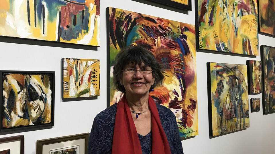 Virginia Pésémapéo Bordeleau pose devant plusieurs de ses tableaux, souriante.