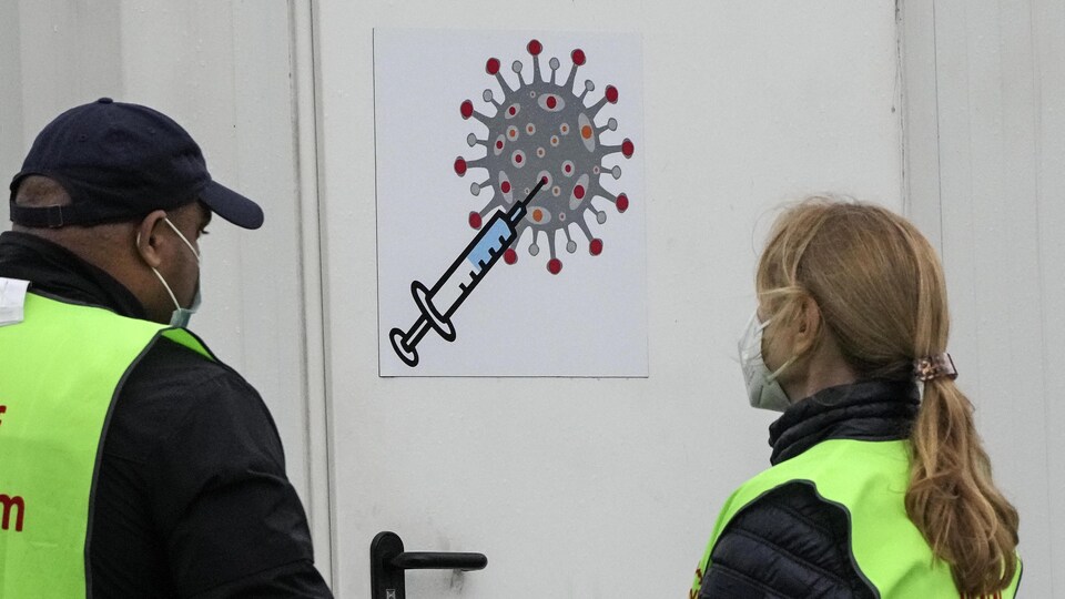 Deux employés d'un centre de vaccination sont de dos. Ils font face à une porte sur laquelle est affiché un pictogramme représentant une seringue piquant un virus.