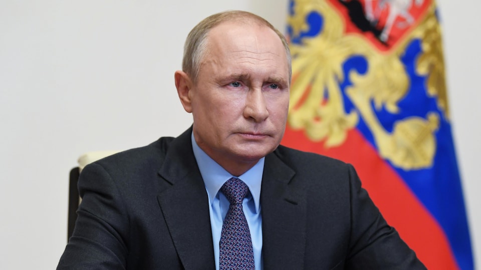 Vladimir Poutine est assis et regarde devant lui, avec un drapeau russe en arrière-plan.