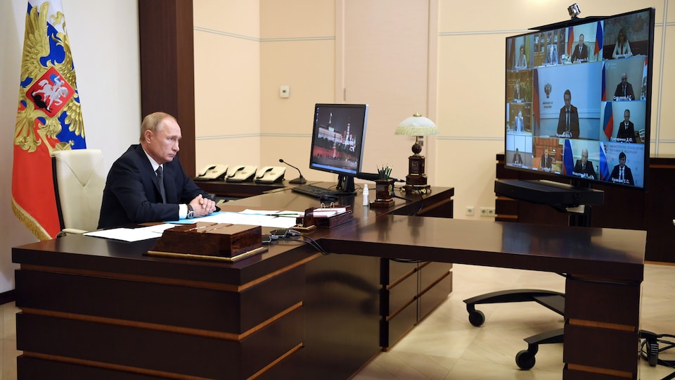 Vladimir Poutine, assis derrière un bureau, regarde un écran où apparaissent les membres de son Cabinet.