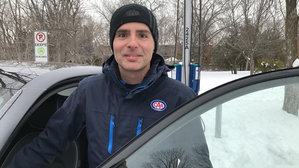 Jesse Caron a garé la voiture électrique qu'il teste ce jour-là devant une borne de recharge dans un parc. Il pose aux côtés du véhicule devant une portière ouverte. 