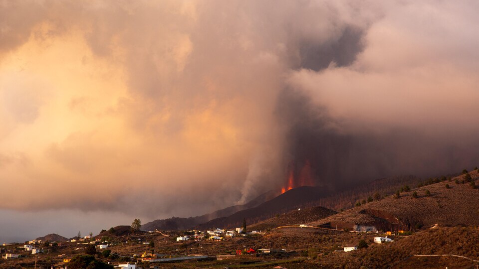 Un volcan en éruption avec des bâtiments en avant-plan. 