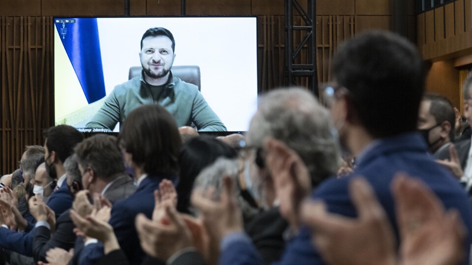 Des gens réunis dans une salle applaudissent tandis que le président ukrainien, dont l'image est diffusée sur grand écran, sourit. 