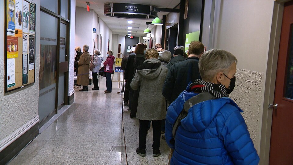 Une dizaine de personnes portant un masque médical attendent dans une file délimitée par une corde.