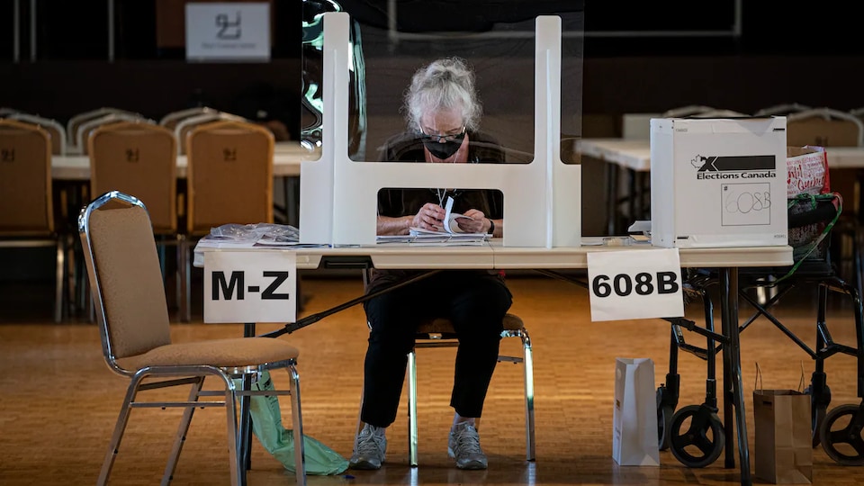 Une femme assise à un bureau de vote, derrière un plexiglas.