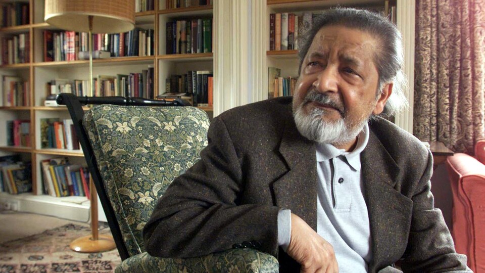 L'auteur britannique V.S. Naipaul, assis sur une chaise dans une bibliothèque.