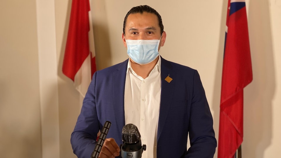 Wab Kinew est debout à un lutrin devant un micro et porte un masque.