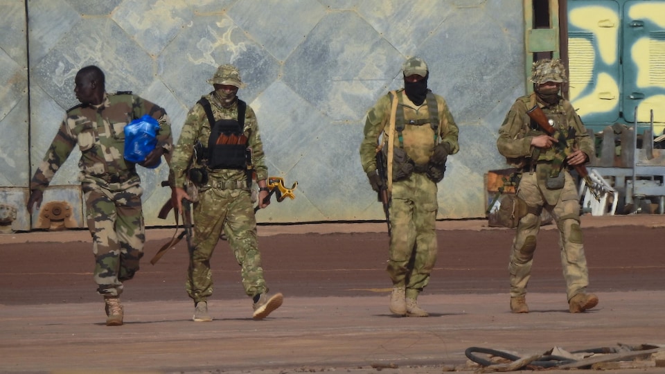 Trois hommes blancs en treillis militaire, le visage masqué, marchent à côté d'un soldat africain.