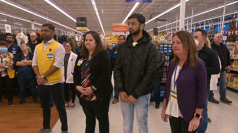 Quatre employé du Walmart, habillé en civil sans un, semblent écouter quelqu’un parler. Ils sont au milieu de leur lieu de travail et des gens, venus magasiner, passent en marchant derrière eux ou bien se sont arrêtés pour écouter ce qui se passe. Il a aussi d’autres employés du Walmart qui écoute. 