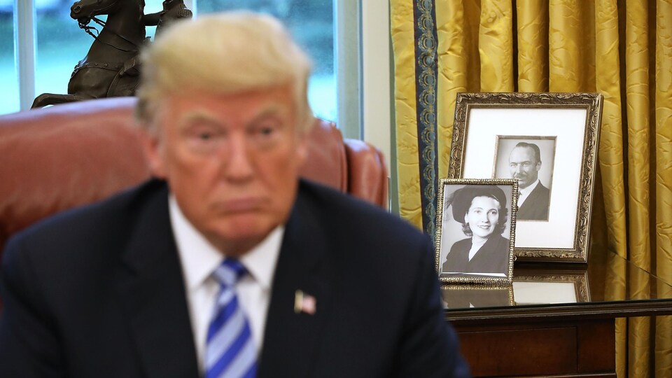 On voit le président Trump à l'avant-plan, mais le point focal porte sur des photographies en noir et blanc de ses parents, sur une table derrière lui. 