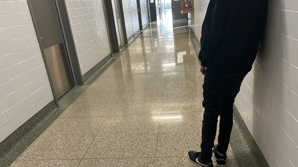 Un jeune appuyé sur un mur de corridor d'une école secondaire.