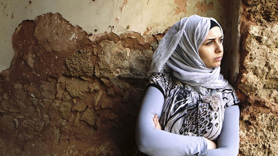 Une femme portant un voile se tient les bras croisés devant un mur.