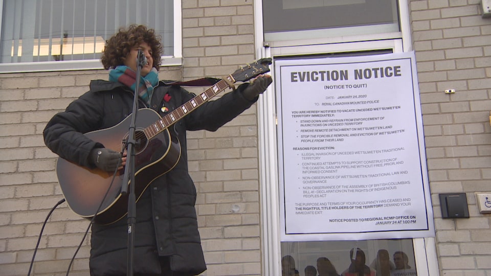 Un chanteur est sur scène en premier plan, devant la porte des bureaux de la GRC sur laquelle un avis d'éviction géant a été placé.