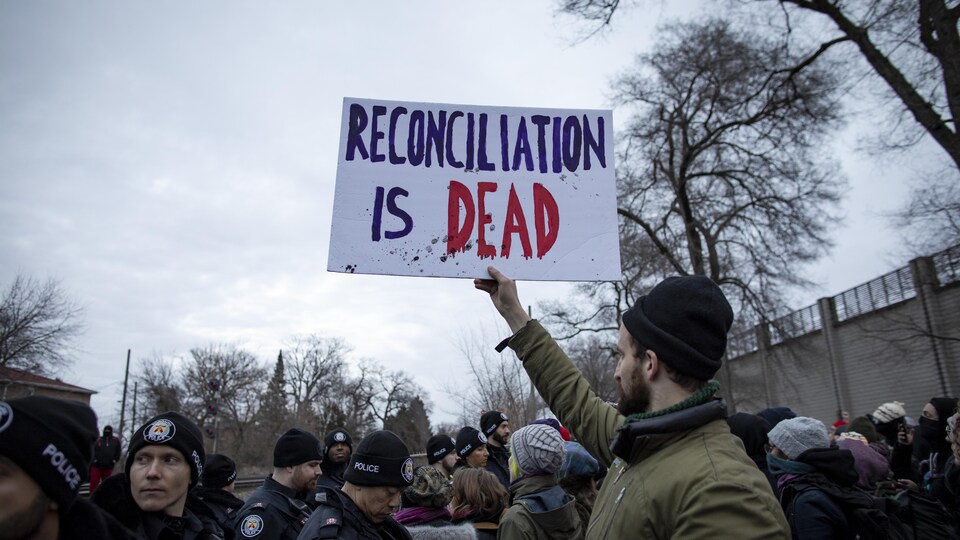 Un membre d'un groupe de manifestants tient une pancarte où il est écrit en anglais : « La réconciliation n'existe plus. » Devant lui, plusieurs policiers surveillent le groupe. 
