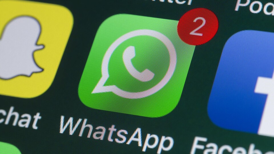 Une photo d'un écran de téléphone sur lequel apparaissent différentes icônes de réseaux sociaux. L'icône de WhatsApp est mise en évidence, au centre, avec une bulle rouge indiquant deux notifications.