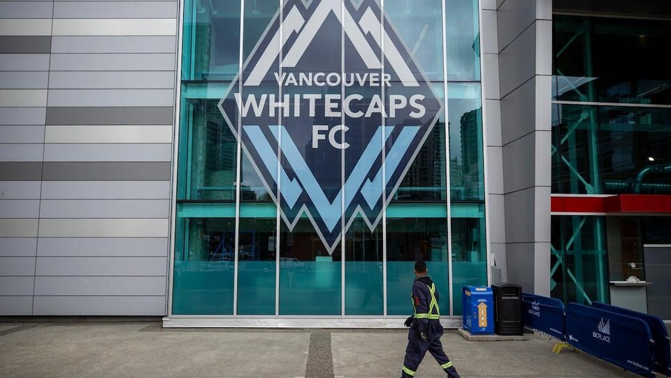Un homme en uniforme de construction marche devant une façade en verre dans laquelle il y a le logo des Whitecaps de Vancouver.