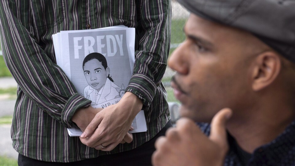Un homme se tient le menton devant une personne tenant des affichettes représentant Fredy Villanueva.