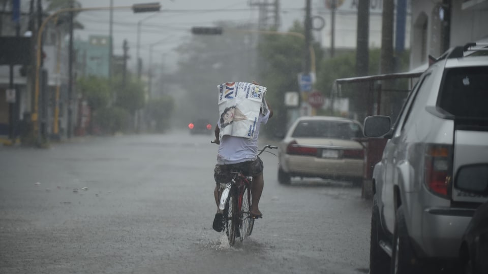 Un homme circule sur un vélo dans une rue recouverte par quelques centimètres d'eau. Le temps est lourd et il pleut.