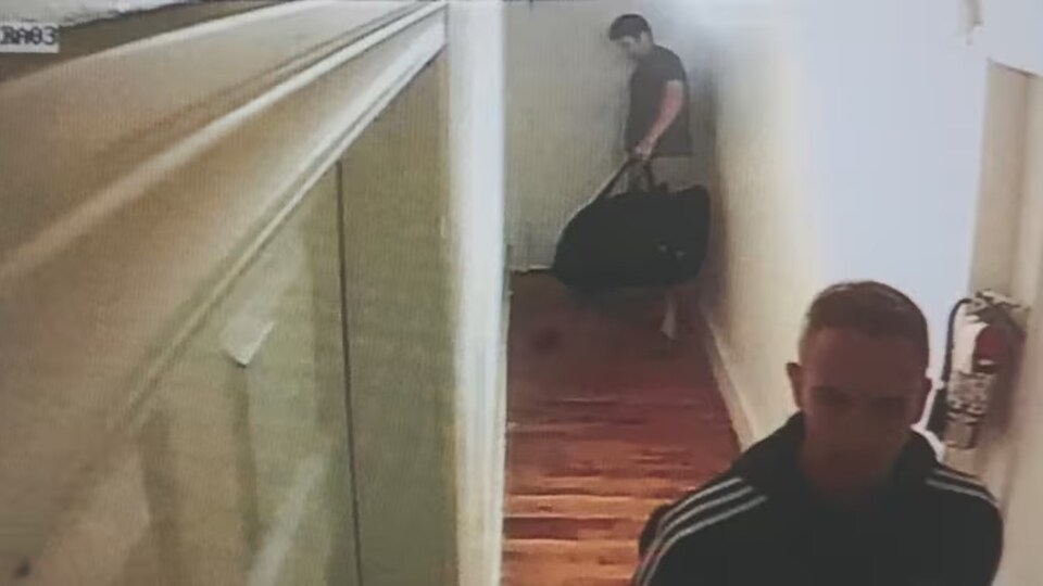 Deux hommes dans un couloir, l'un porte un sac.